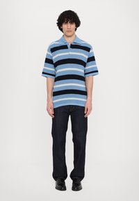 Kortermet polo-shirt i lyseblå med marineblå og hvite horisontale striper, kombinert med mørke jeans og svarte sko. Modellen står rett opp.