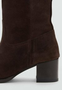 Brązowy zamszowy botek na blokowym obcasie, z widocznymi szwami i prostą, czystą sylwetką. Gładka, matowa tekstura w całości.