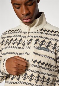 Pull en polaire crème avec des motifs géométriques noirs et beiges, col zippé à mi-hauteur et poche avant avec la marque Columbia.