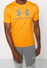 Oranžové tričko s krátkým rukávem a šedým logem "Under Armour" na přední straně. Materiál z bavlněné směsi, kulatý výstřih a volný střih.