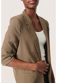 Marynarka w kolorze oliwkowej zieleni z wyciętym klapą, zapięciem na jeden guzik i lekko podwiniętymi rękawami. Gładka faktura i detal z przednią kieszenią.