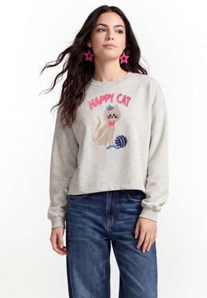 Donna che indossa una felpa grigio chiaro con gatto testurizzato, gomitolo di lana e testo rosa "HAPPY CAT", abbinata a jeans blu e orecchini a forma di stella rosa.