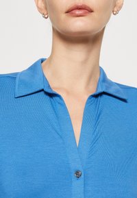 Blue collared shirt made of smooth fabric, featuring a button closure and a V-neck design. The collar lies flat with neat stitching.