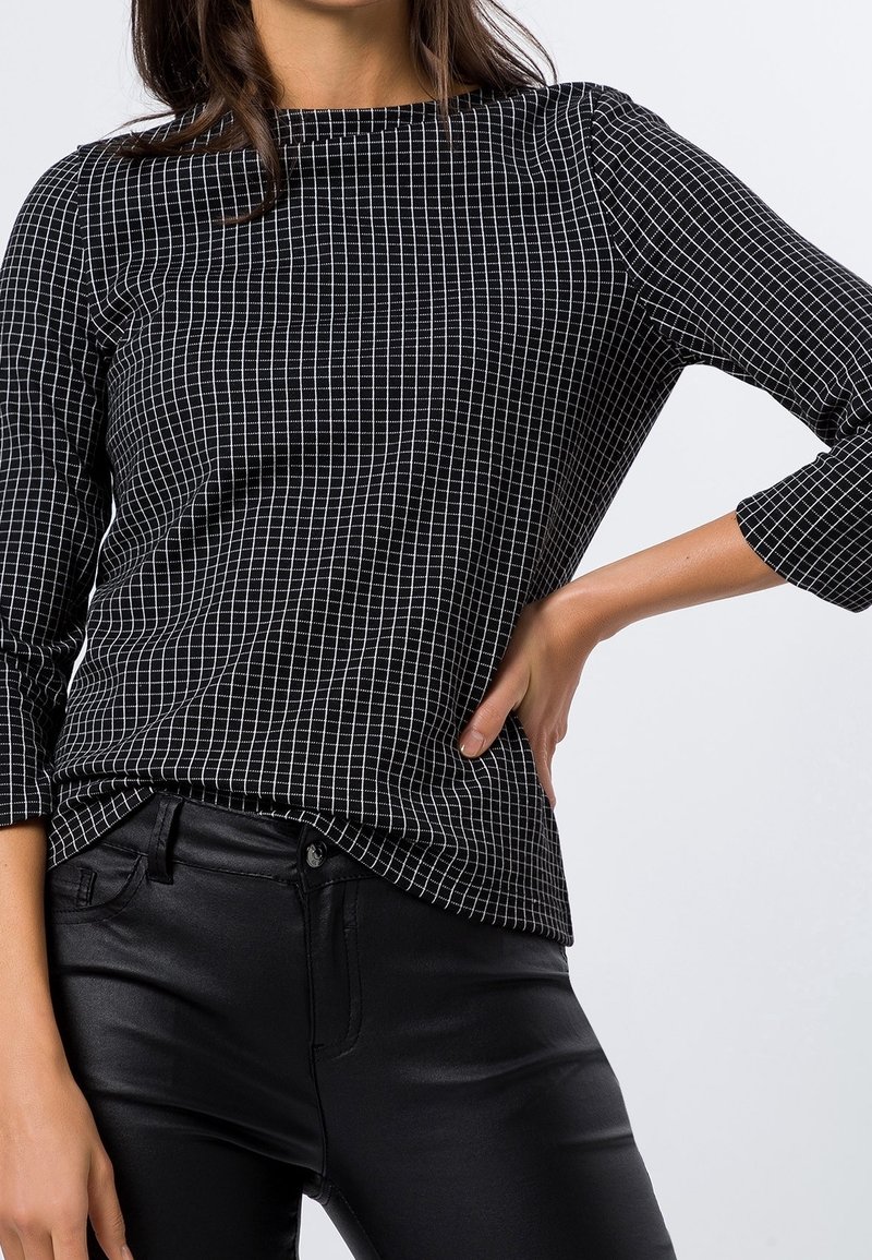 Femme portant un haut noir à manches longues avec un motif quadrillé blanc et un pantalon noir ajusté, posant avec une main sur la hanche.