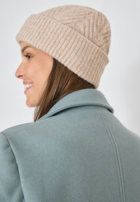 Beige gebreide beanie met ribbelstructuur en gevlochten patroon, gedragen met een blauwe wollen jas met een geknipte kraag en een gladde afwerking.