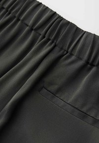 Schwarze Stoffhose mit elastischem Bund und einer einzelnen Leistentasche auf der Rückseite, in Nahaufnahme gezeigt.