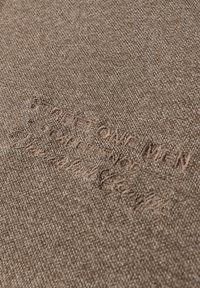 Brauner Stoff mit einer subtilen Heather-Struktur und geprägten Branding-Details. Das Material ist weich, wahrscheinlich eine Baumwollmischung.