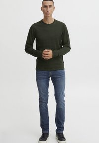 Chemise à manches longues vert foncé avec un col rond, associée à un jean bleu délavé et des baskets noires. Design simple sans motifs visibles.