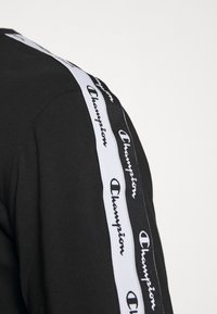 Schwarzer Stoff mit zwei parallelen Champion-Logo-Streifen, einer weißen und einer schwarzen, die vertikal entlang eines Ärmels verlaufen.