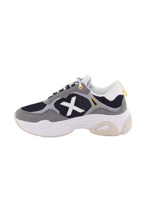 Las zapatillas presentan una combinación de parte superior de gamuza gris y malla, con acentos en negro, suela blanca y detalles en amarillo. El diseño incluye un gran logo blanco en el costado.