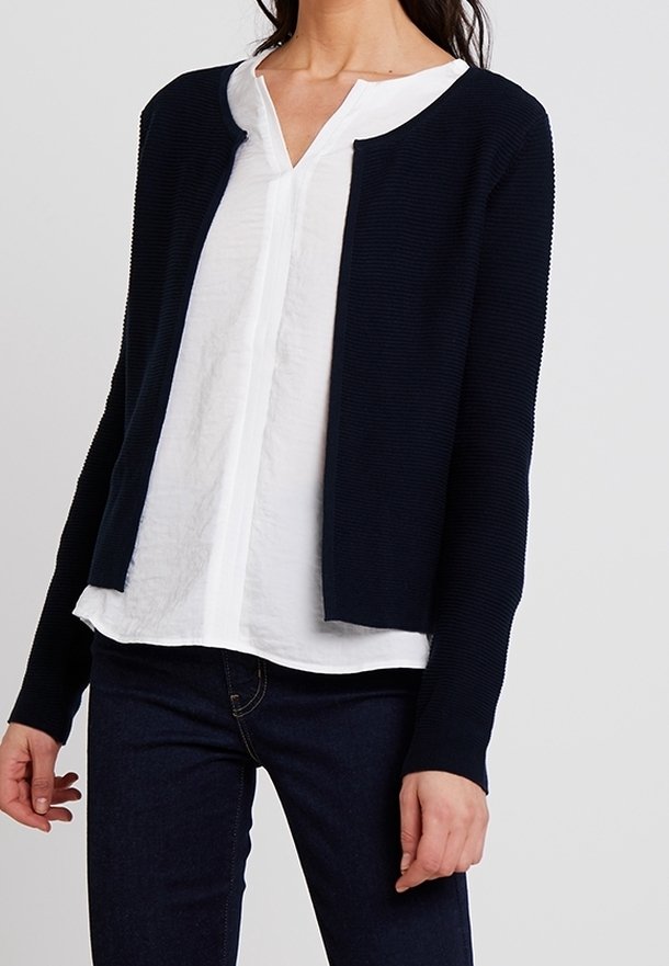 Femme portant un chemisier blanc à col en V sous un cardigan bleu marine foncé ouvert sur l'avant avec des manches longues et un pantalon foncé.