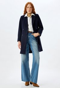 Abrigo de marino de doble botonadura, camisa clara con cuello, suéter de cuello alto color beige, jeans de mezclilla claros de pierna ancha, botas marrones de tacón, accesorios mínimos.