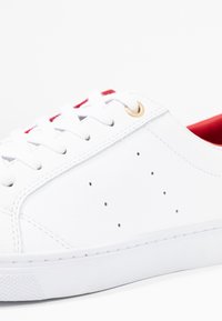 Zapatilla de cuero blanca con interior rojo, ojetes dorados y detalles perforados en la parte superior. Suela de goma con agarre texturizado.