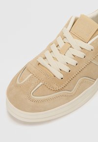 Sneaker in pelle scamosciata beige con superficie texturizzata, dotata di lacci crema, cuciture a contrasto e una etichetta del logo sulla linguetta.