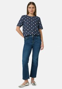 Navy-blau gemusterte Kurzarmbluse mit hellblauen und weißen Rauten; kombiniert mit geraden blauen Jeans und silbernen Ballerinas.
