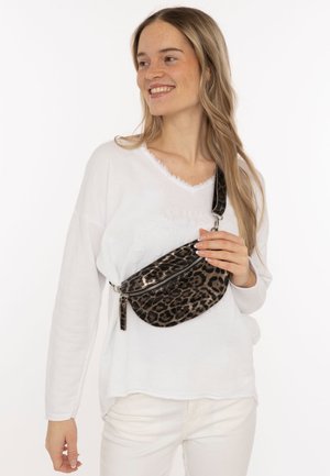 Lächelnde Frau in weißem Outfit hält eine Umhängetasche mit Leopardenmuster und Reißverschluss, mit dem Riemen über der Schulter, und schaut zur Seite.