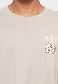 Lys beige t-shirt med rund hals. Har et sort isterning grafik og Adidas trefoil-logo i hvid på brystet. Blødt bomuldsstof.