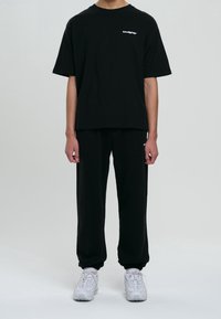 Svart t-shirt med kort ärm och en vit logotyp på bröstet, i kombination med matchande lösa svarta joggers och grå sneakers.