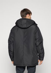 Giacca a vento nera con cappuccio, caratterizzata da una texture liscia, vestibilità over size e polsini elasticizzati. La vista posteriore mostra un design elegante e minimal.