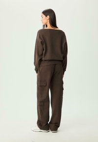 Felpa marrone oversize con scollo ampio, abbinata a pantaloni cargo coordinati con molteplici tasche laterali. Sneakers bianche completano l'outfit.