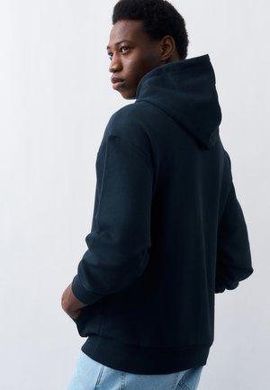 Jaunas vyras dėvi tamsiai mėlynę džemperį su gobtuvu ir šviesius džinsus, stovintis su rankomis kišenėse, žiūrintis į šoną priešais paprastą foną.