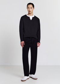 Schwarzes Langarmshirt mit V-Ausschnitt und drei Knöpfen, kombiniert mit schwarzen Hosen. Weiße Sneaker mit hellen Akzenten.