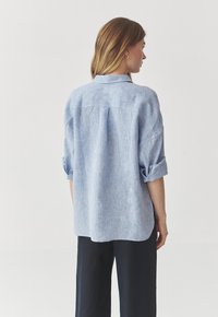 Camisa de un azul claro, con apariencia de lino, mangas arremangadas, una costura en la yugo trasero y un dobladillo redondeado. La tela tiene una textura sutil.