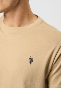 Beige bomulds T-shirt med korte ærmer, prydet med et navy broderet logo på venstre bryst. Glat tekstur, klassisk rundhalset design.