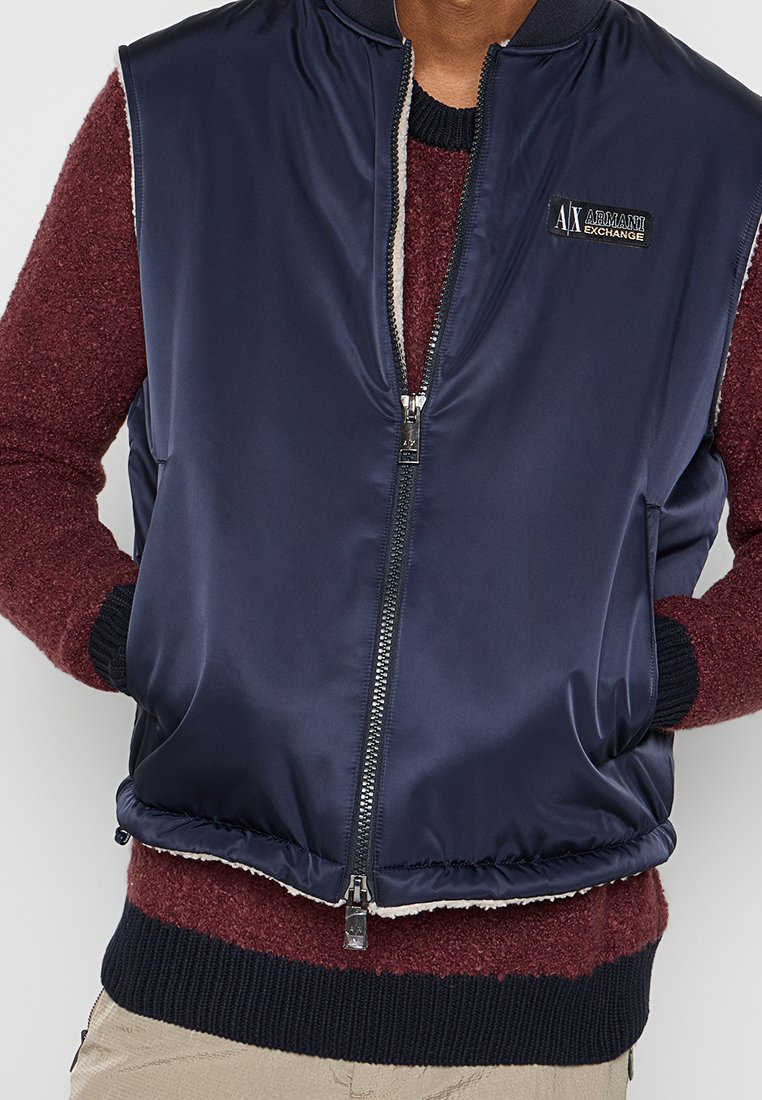 Gilet imbottito blu navy con zip frontale, colletto a costine e una patch con logo. Presenta tasche laterali e una morbida fodera interna.