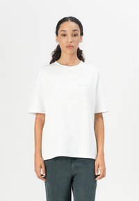 White cotton t-shirt with short sleeves, loose fit, crew neckline, and light blue printed text on the front. Paired with dark denim pants.