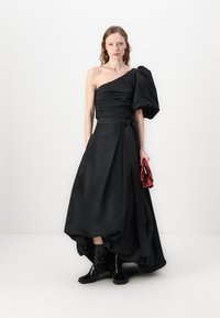 Abito nero a un spalla con manica sovradimensionata drappeggiata; vita arricciata e gonna fluida. La modella tiene una piccola borsa rossa e indossa stivali neri.