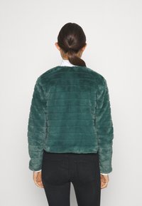Beskuren teal-faux pälsjacka med horisontell sömnad och rundad halsringning. Matchad med svarta jeans som visar en jämn textur.