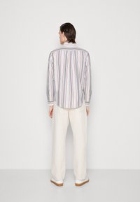 Camisa a rayas de tejido suave con mangas largas, con rayas estrechas en azul, rosa y amarillo sobre un fondo blanco. Combinada con pantalones claros.