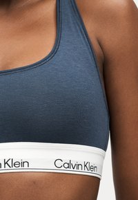 Κοντινό πλάνο ενός ατόμου που φοράει αθλητικό σουτιέν Calvin Klein σε σκούρο μπλε χρώμα με λευκή ελαστική ταινία όπου αναγράφεται το όνομα της μάρκας με μαύρο κείμενο.
