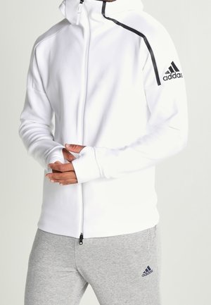 Osoba w białej bluzie z kapturem Adidas zapinanej na zamek z czarnym paskiem na ramieniu i szarych spodniach dresowych Adidas, dłonie splecione na wysokości talii.