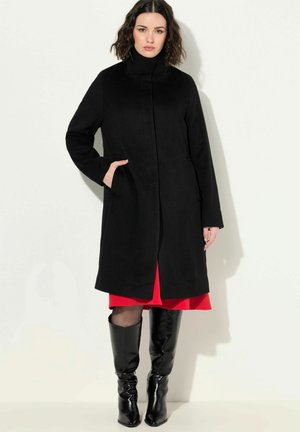 Schwarzer Mantel mit hohem Kragen, seitlichen Taschen und geradem Schnitt. Über einem roten Kleid getragen, kombiniert mit schwarzen kniehohen Stiefeln.