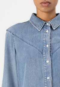 Gros plan sur une personne portant une chemise en denim bleu clair avec des boutons blancs et un col pointu sur un fond blanc.