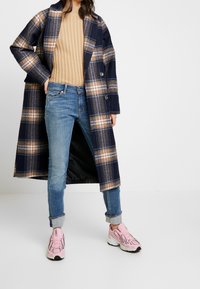 Femme portant un long manteau à carreaux bleu marine, un pull beige à côtes, un jean bleu retroussé et des baskets roses, debout devant un fond blanc.