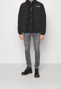 Giacca a vento nera con colletto alto e trapuntatura orizzontale, abbinata a una felpa nera con zip, jeans grigi strappati e stivali neri.