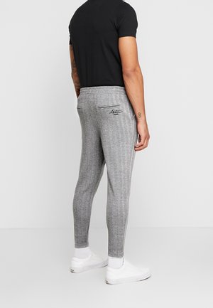 Mand iført slim-fit grå ternede bukser med sort skrift på baglommen, sort t-shirt, hvide sokker og hvide sneakers, stående sideværts.
