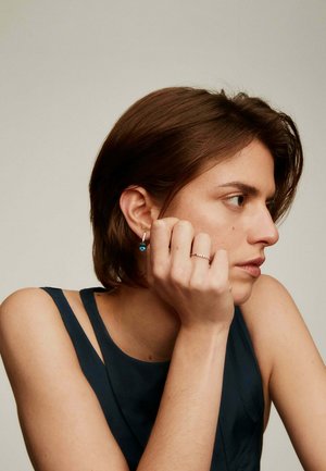 Pendientes de plata con una piedra preciosa azul, llevados por una persona con cabello corto castaño, que viste una blusa sin mangas oscura, con la mano apoyada en la barbilla.