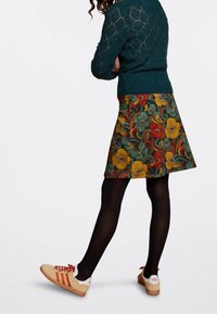 Hame floral-kuvioilla, jossa on vihreitä, keltaisia ja punaisia kukkia tummalla taustalla; yhdistettynä mustiin sukkahousuihin ja beigneihin lenkkareihin, joissa on punaisia yksityiskohtia.