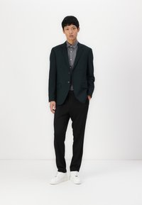 Dunkelgrüner Blazer über einem grauen Hemd, kombiniert mit schwarzen Hosen und weißen Sneakers. Der Blazer hat zwei Knöpfe und eine taillierte Passform.