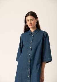 Denim obleka v temno modri barvi s ovratnikom, 3/4 rokavi in gumbi po sprednji strani. Mehka tekstura in sproščen kroj.