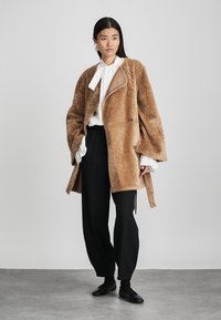 Ruskea shearling-takki, jossa on suuri kaulus, tan-sävyinen yksityiskohta ja kuusi pyöreää nappia. Yhdistetty mustiin housuihin ja valkoiseen paitaan.