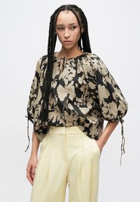 Luźna bluzka w czarno-beżowy kwiatowy wzór, z okrągłym dekoltem i regulowanymi sznureczkami, zestawiona z żółtymi, wysokim stanem spodniami.
