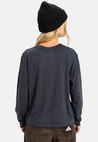 Mørkegrå langermet skjorte med rund hals, avslappet passform og et merke ved kanten; kombinert med brune cordbukser og en svart lue.