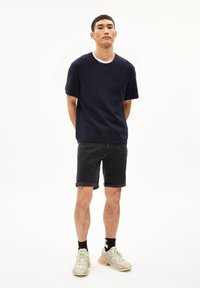 Marineblauwe korte mouwen crewneck trui, gecombineerd met donkere denim shorts. Model draagt lichte sneakers met pastelaccenten en zwarte sokken.