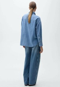 Denimhemd mit lässigem Schnitt, langen Ärmeln und klassischem Kragen, kombiniert mit weit geschnittenen Denim-Hosen. Hellblau mit minimalen Details.