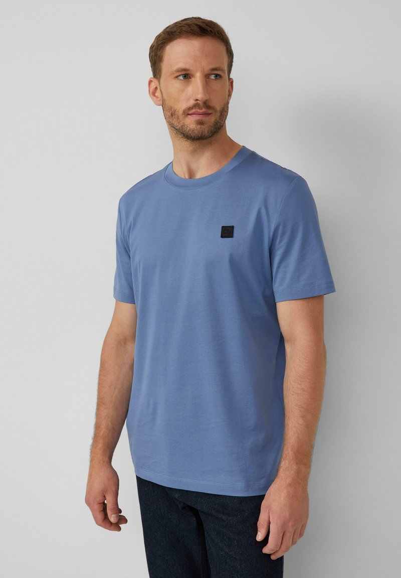 Blauwe katoenen T-shirt met een ronde hals, korte mouwen en een klein zwart vierkant logo op de linker borst. De stof lijkt glad en licht van gewicht.